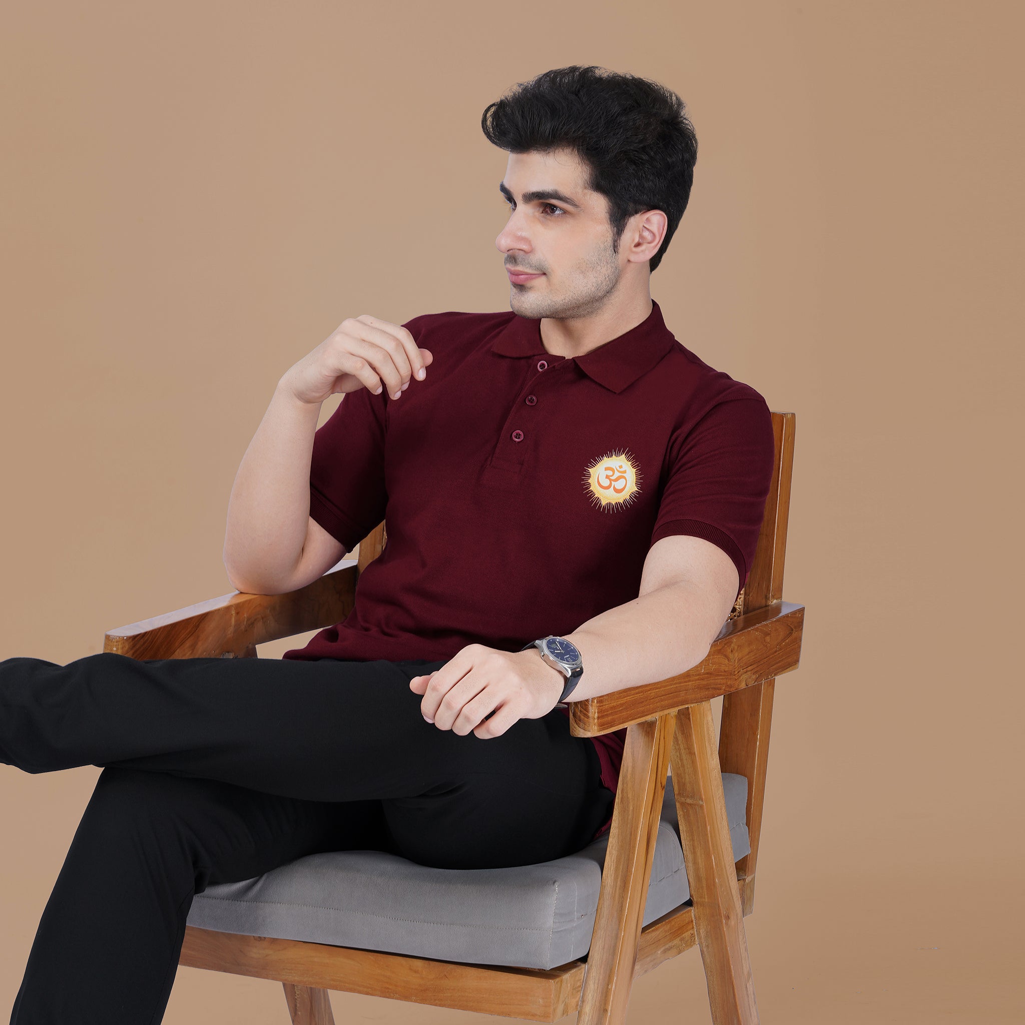 Man wearing the maroon Parambhakt Om Polo T-shirt while sitting on a wooden chair, showcasing a modern spiritual lifestyle look.